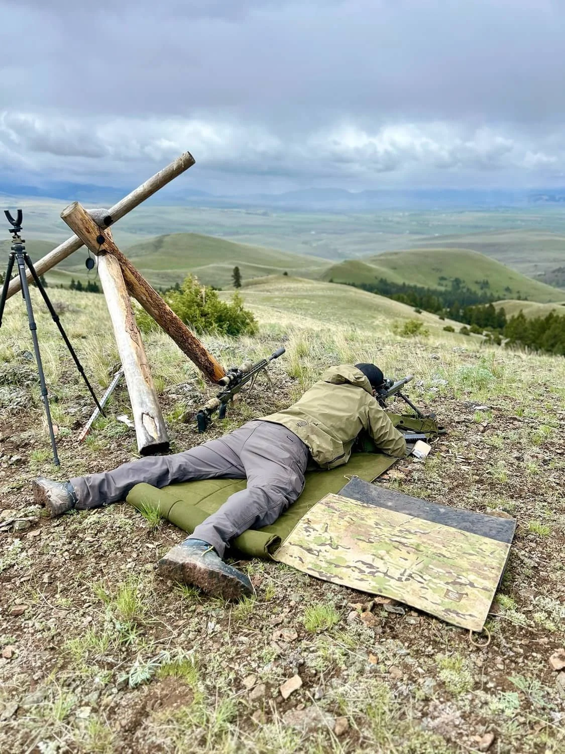 A person lying prone on a mat with a rifle, aiming through a scope in a hilly outdoor landscape with cloudy sky.