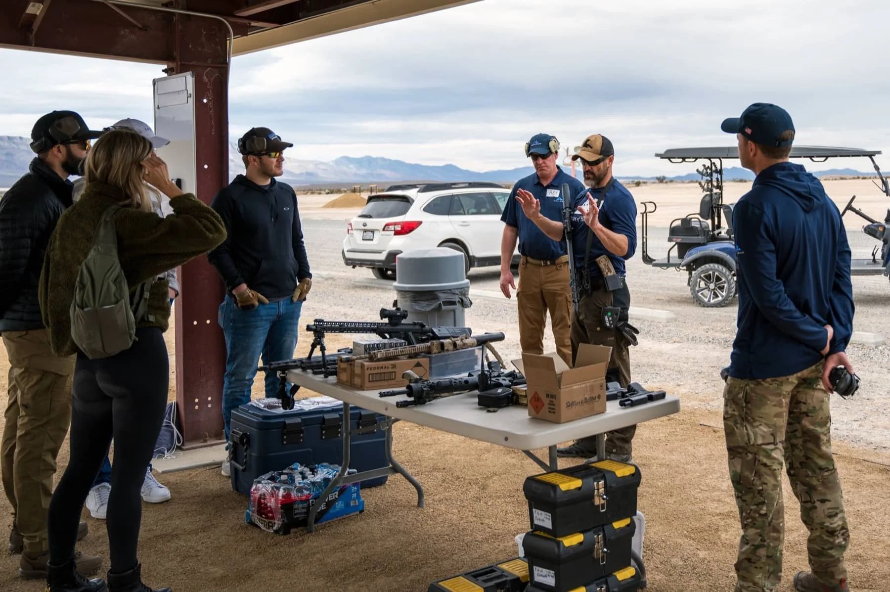 Group of seven people, including officers and civilians, standing around a table with firearms and equipment at an outdoor shooting range in a desert. There are cars and golf carts in the background under a cloudy sky.