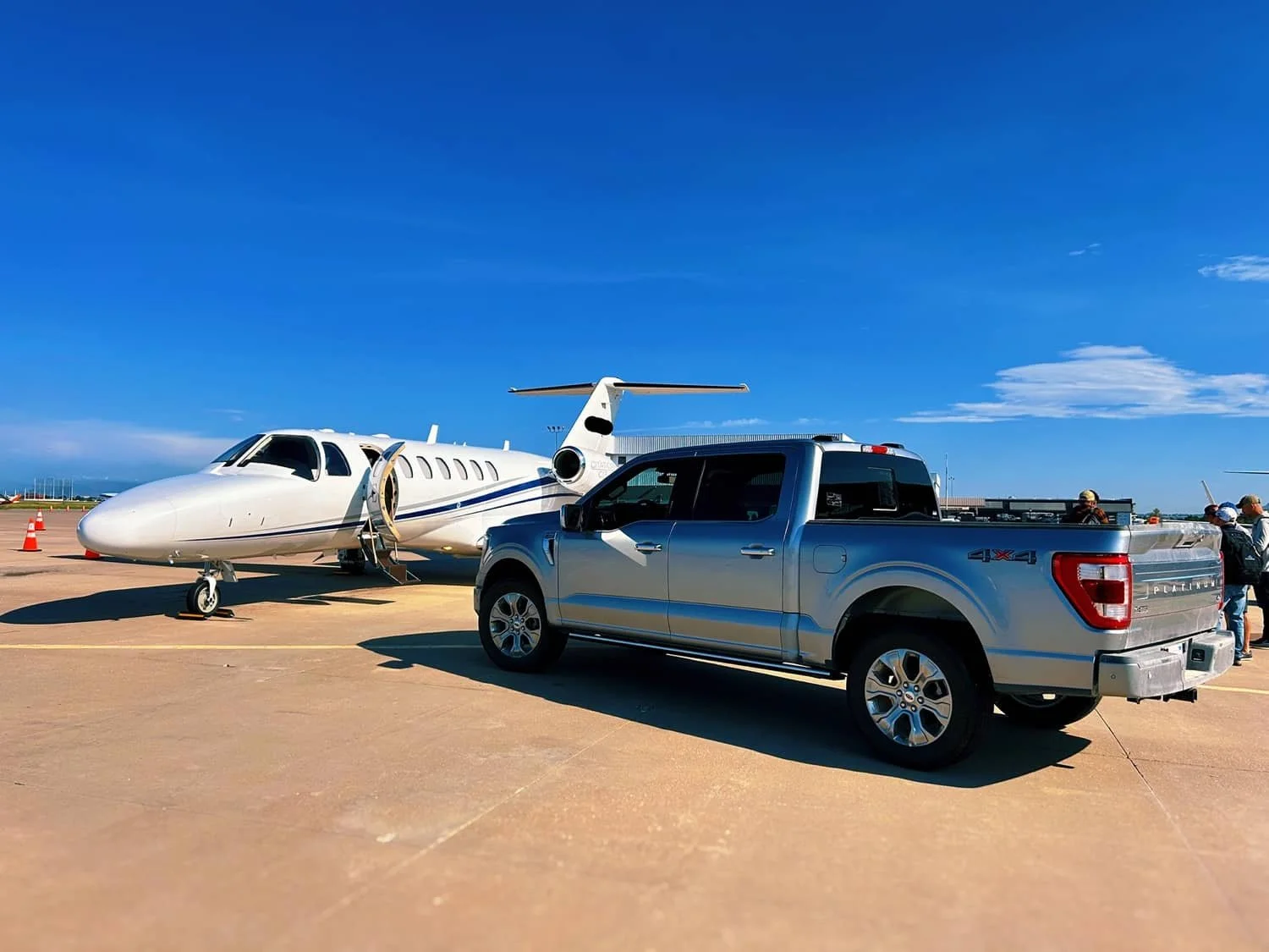 A small white private jet with its door open parked on the tarmac next to a silver pickup truck, with a group of people nearby and an open sky above.