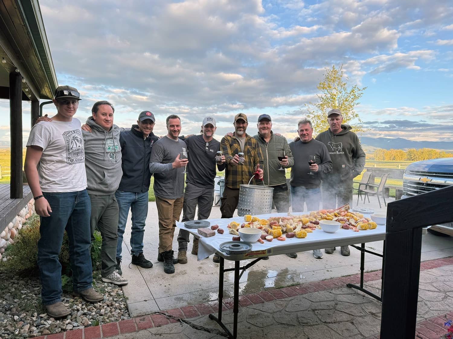A group of nine men standing outside at a barbecue with food on a table, holding drinks, with a scenic landscape and cloudy sky in the background.