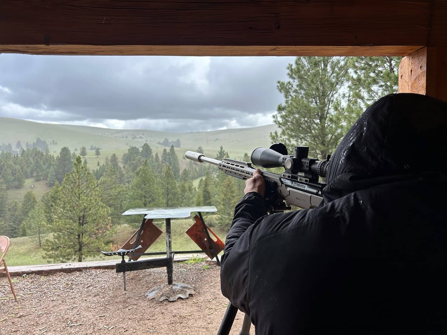 Person in black jacket aiming a rifle with a scope from a wooden shooting perch at a scenic outdoor range with trees and cloudy sky in the background.