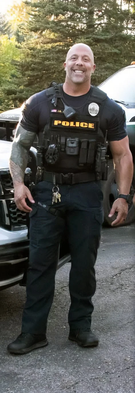 A smiling police officer standing outdoors next to a police vehicle, wearing a tactical vest, black uniform, and carrying keys.