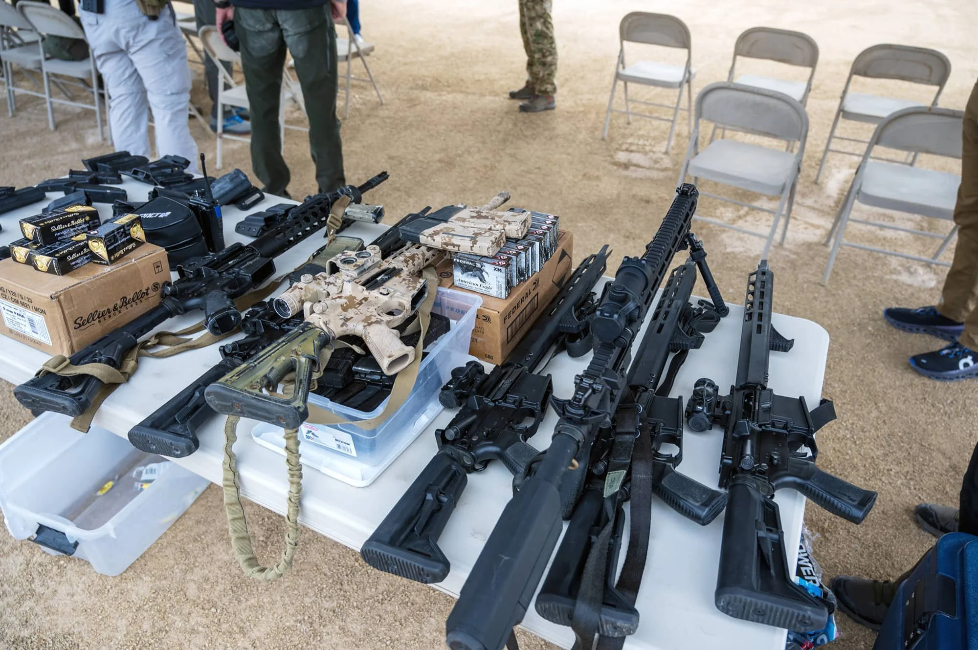 Table displaying various firearms including rifles, handguns, and ammunition, with chairs and people standing around in the background.
