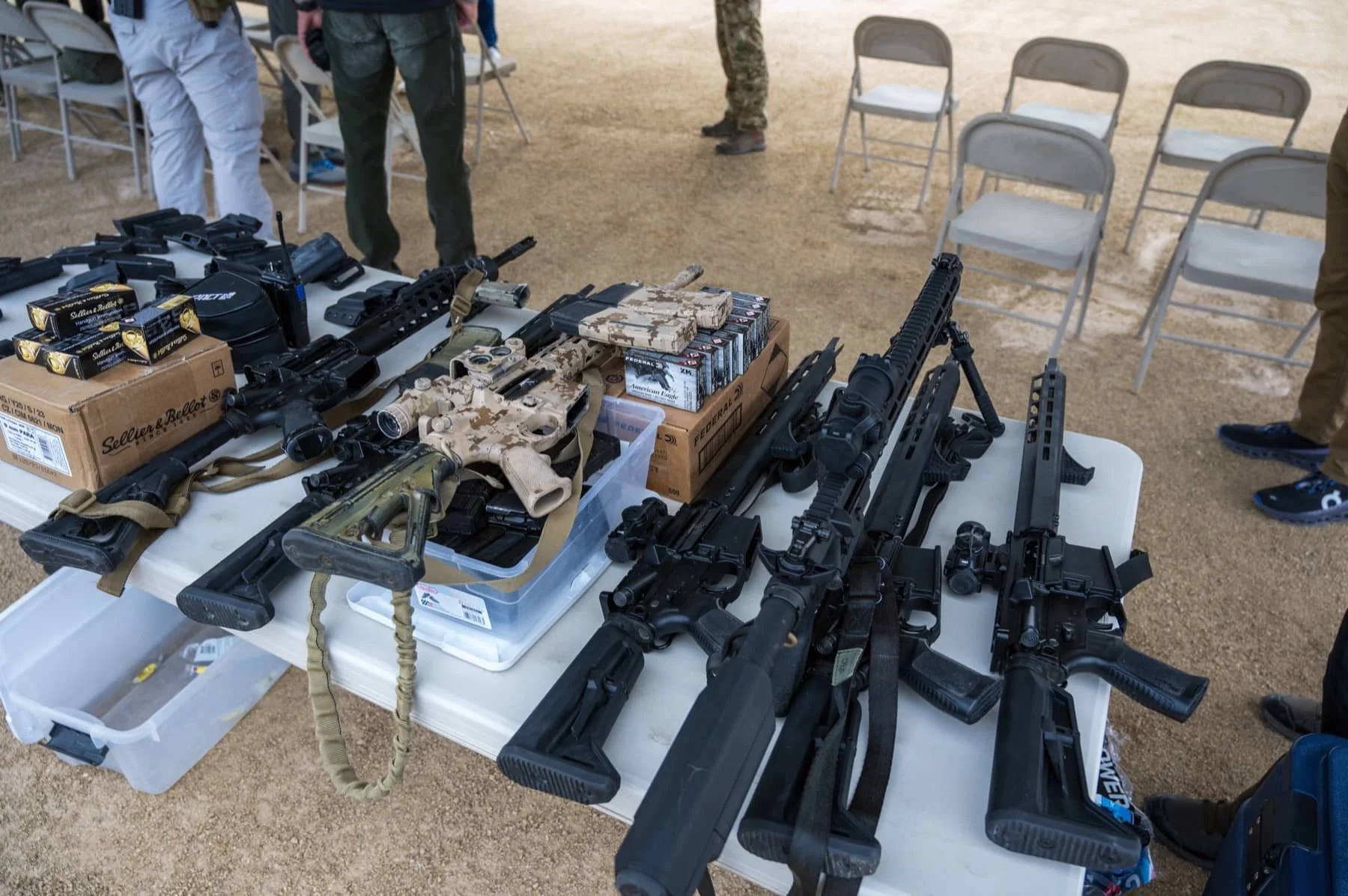 Table displaying various firearms, including rifles and handguns, along with ammunition and accessories, at an outdoor event with folding chairs and people in the background.