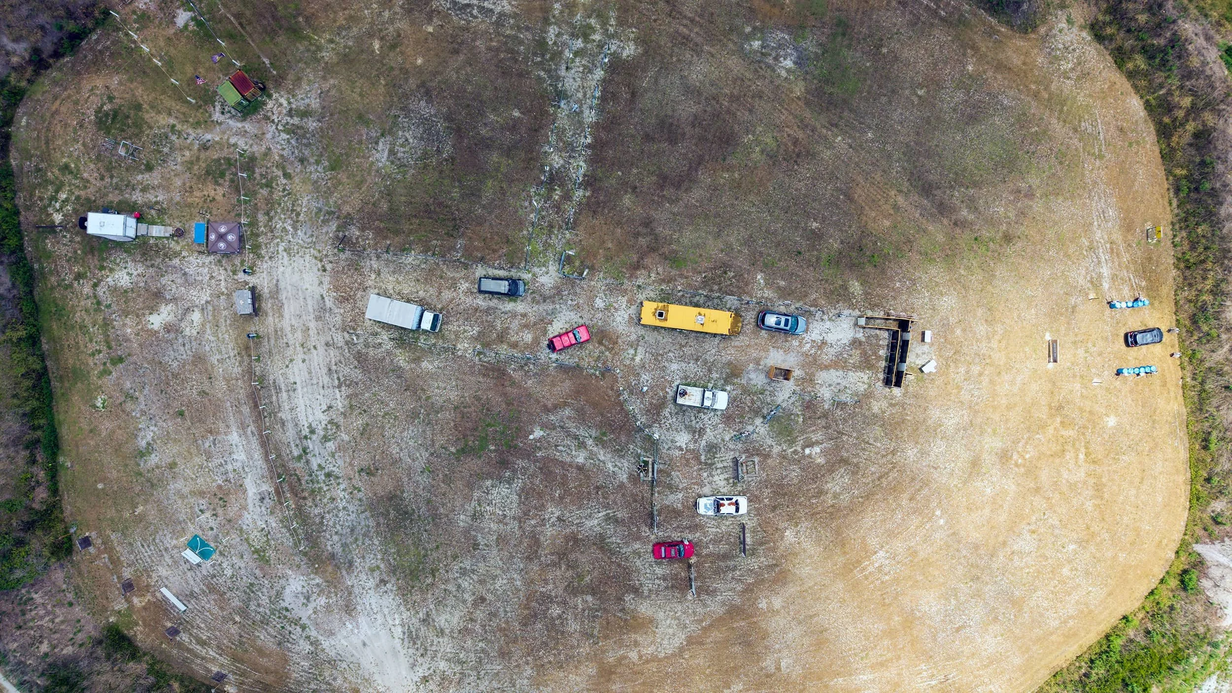 An aerial view of a large, open grassy field with various parked vehicles, including trucks, cars, and trailers, scattered across the area. There are some fenced sections and equipment, along with patches of bare ground amid the grass.