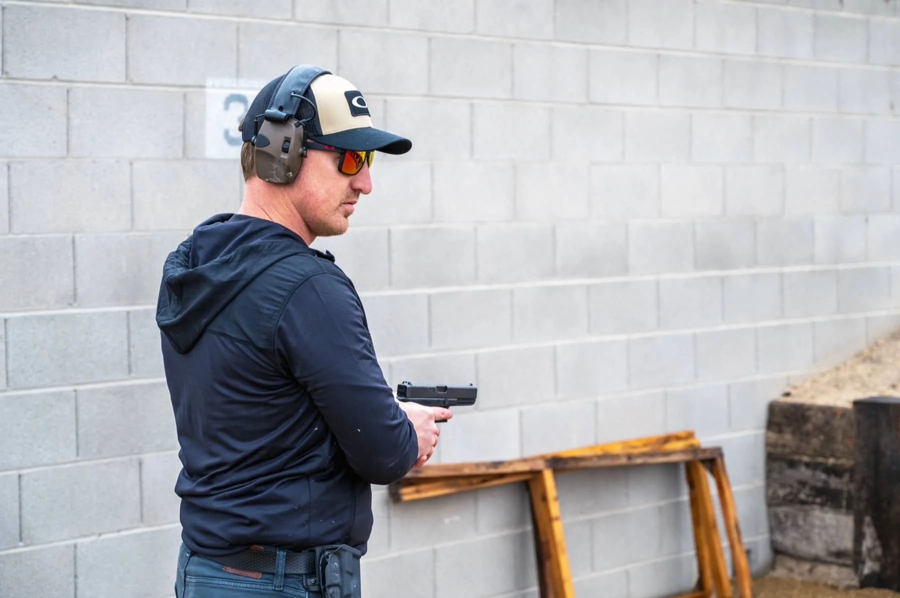 A man wearing sunglasses, a baseball cap, and ear protection holding a handgun at a shooting range.