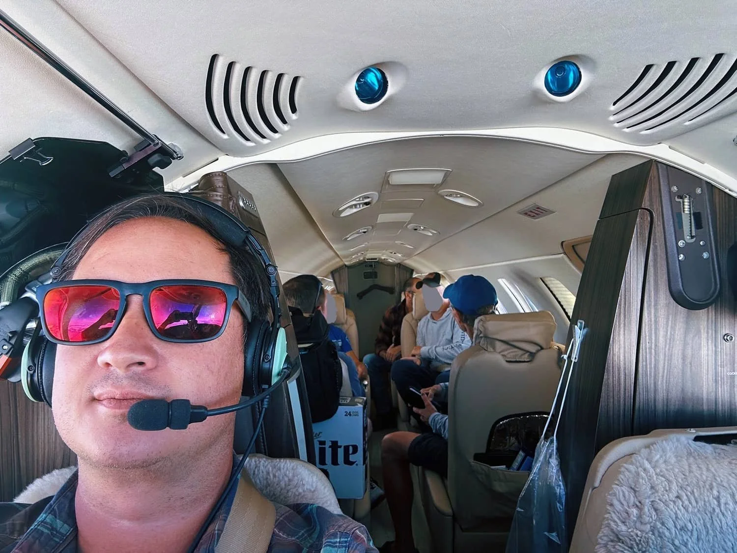 Inside a small airplane cabin with multiple passengers seated. The person in the foreground is wearing sunglasses, a headset, and a plaid shirt. The cabin has wooden accents, overhead speakers, and personal items on the seats.