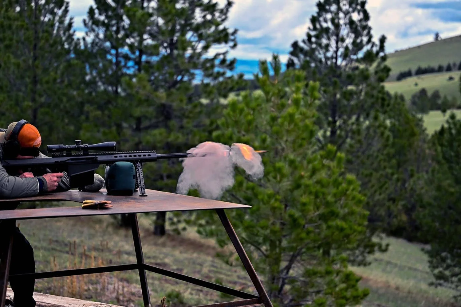 A person firing a sniper rifle from a mounted platform outdoors, surrounded by trees and hills in the background.