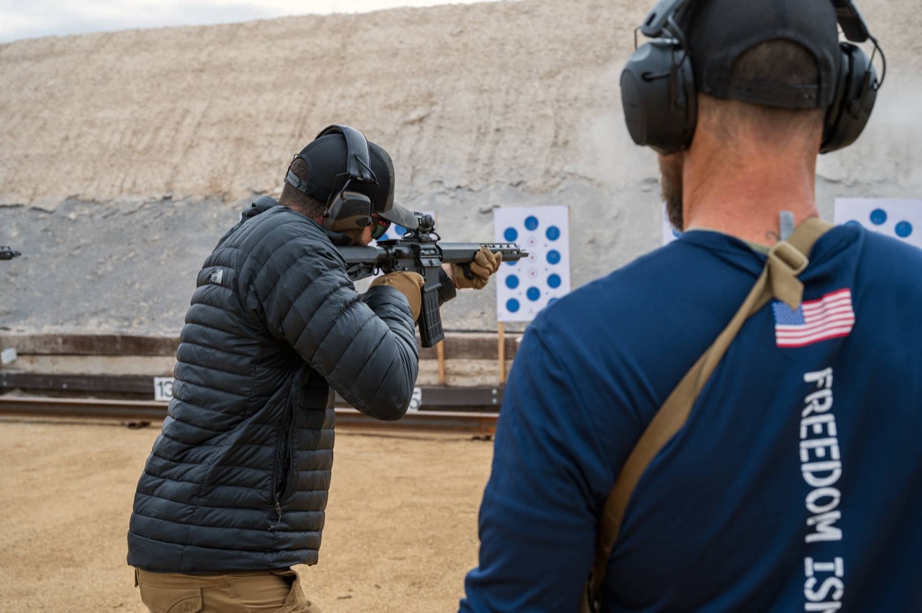 Two men at a shooting range, one aiming a rifle and the other observing, wearing hearing protection, hats, and warm clothing.