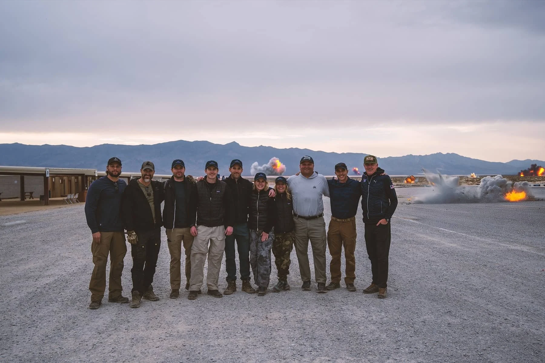 Ten people standing in a line outdoors with explosions and smoke in the background, suggesting a military or testing site.