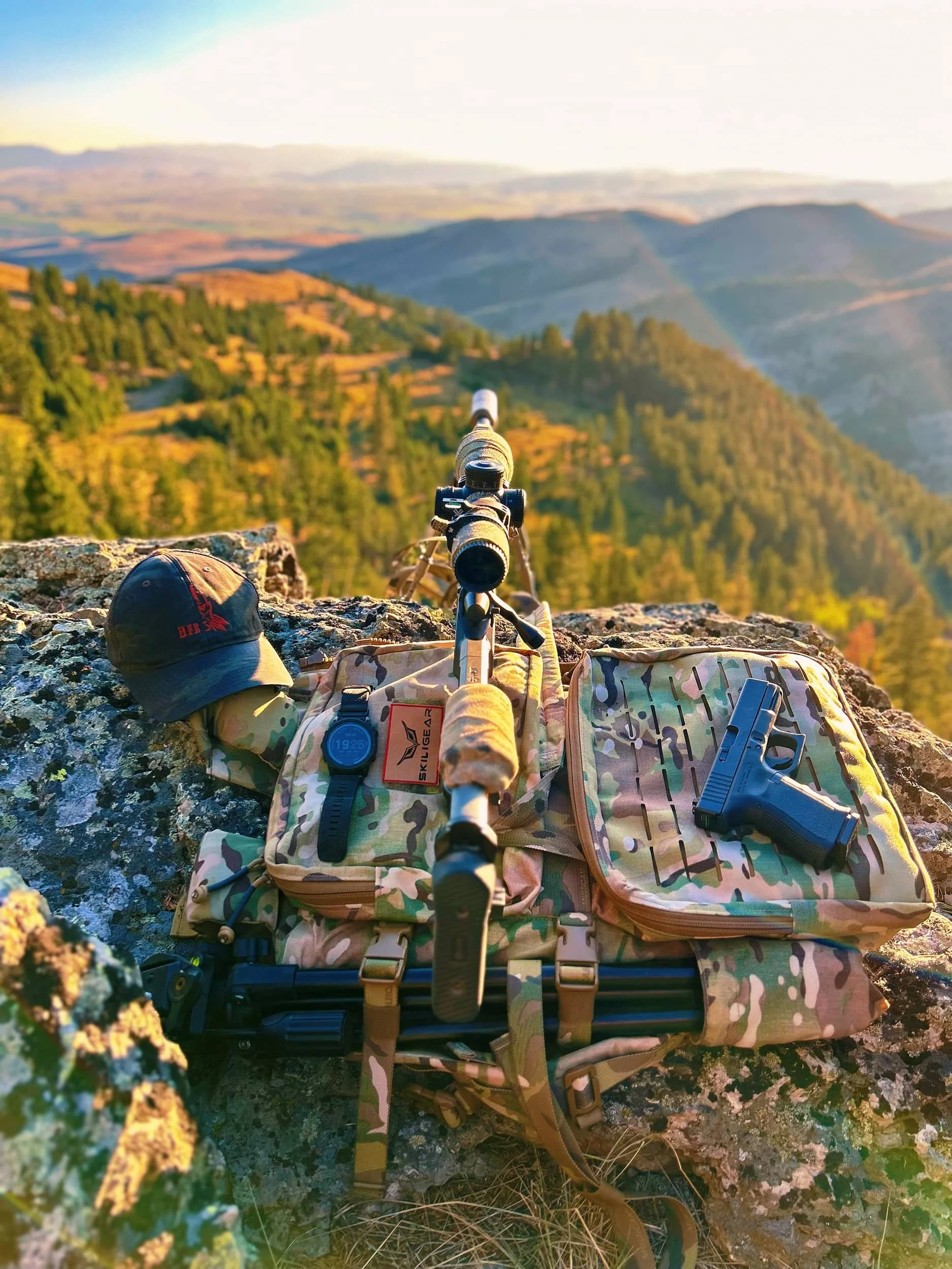 Military camouflage backpack, pistol, baseball cap, and rifle on a rocky ledge overlooking a forested mountain landscape at sunset.