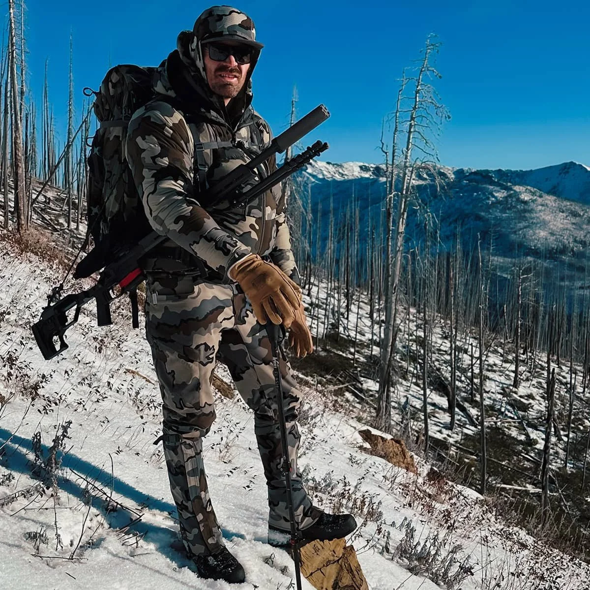 A man in camouflage military gear wearing sunglasses and a helmet, smiling, holding a rifle with a scope, against a clear blue sky with snow-covered mountains in the background.