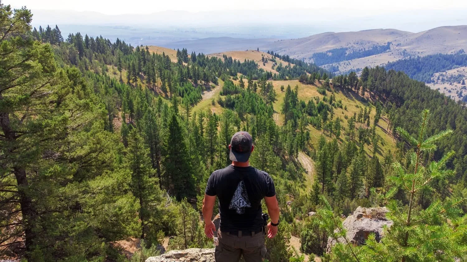 A man standing on a rock ledge overlooking a lush, green forested mountain landscape.