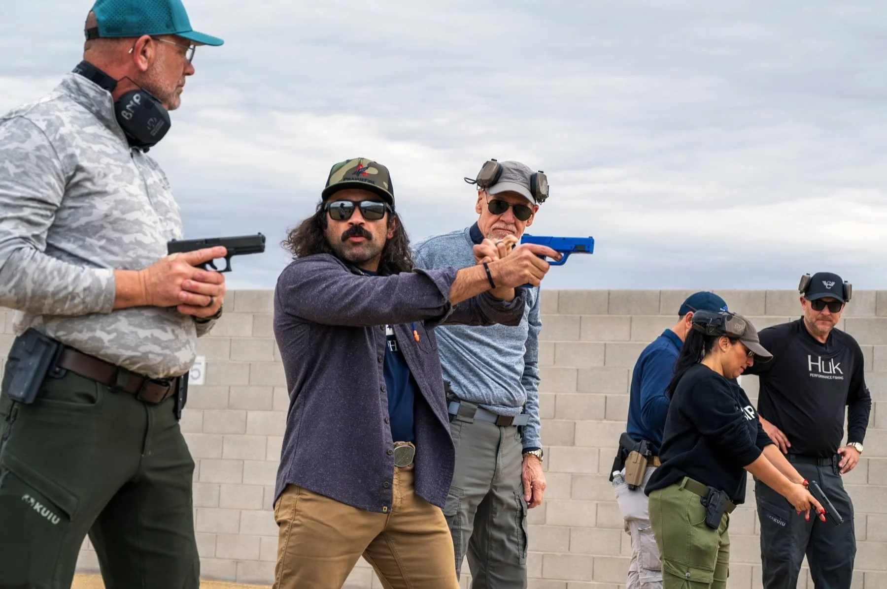 Group of people participating in a shooting activity outdoors, with a concrete wall and cloudy sky in the background. One person is aiming a blue pistol, while others hold firearms and wear safety gear.