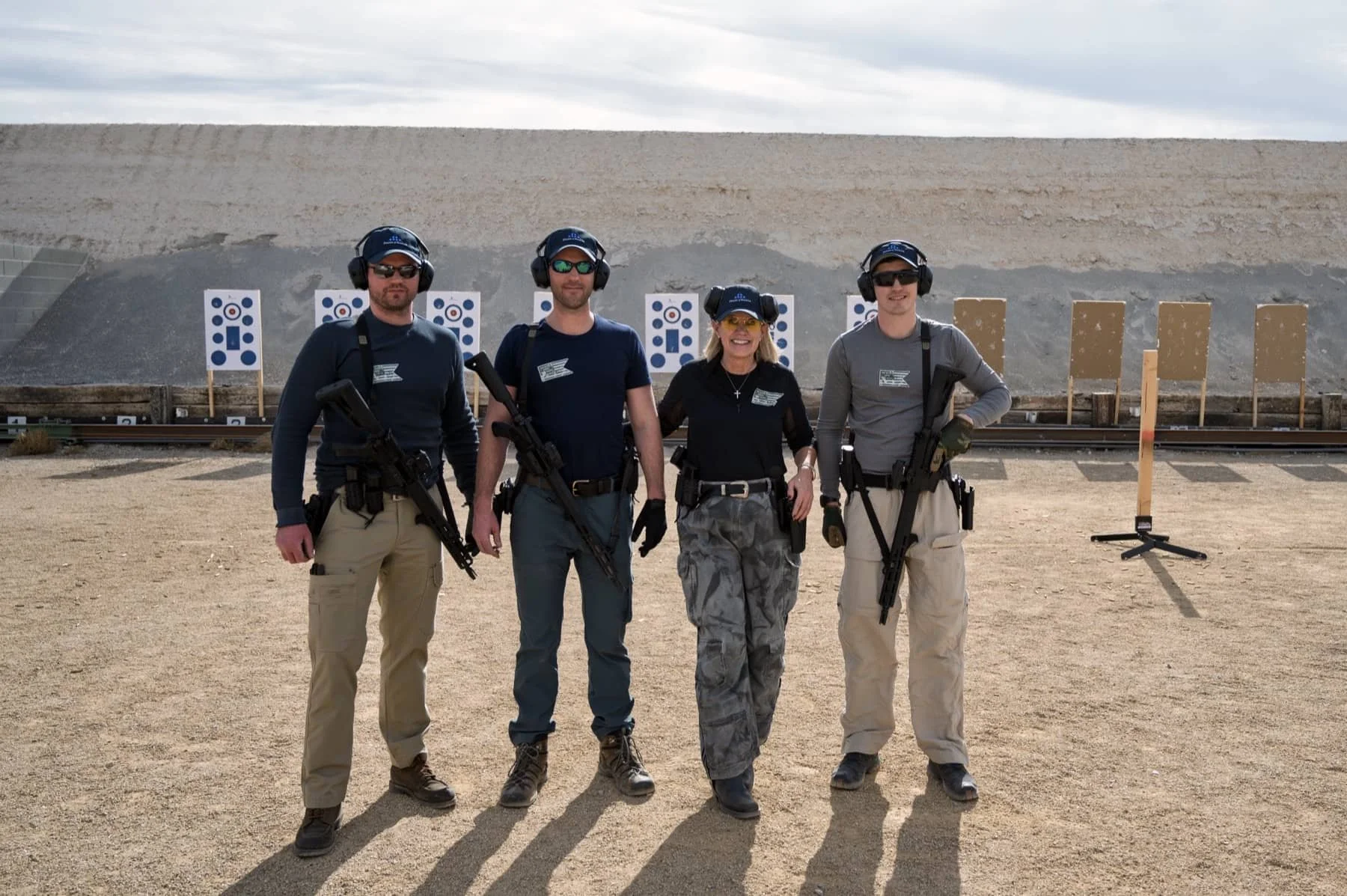 Four individuals at a shooting range, standing side by side with firearms, wearing ear protection, in front of target boards and a sandy backdrop.
