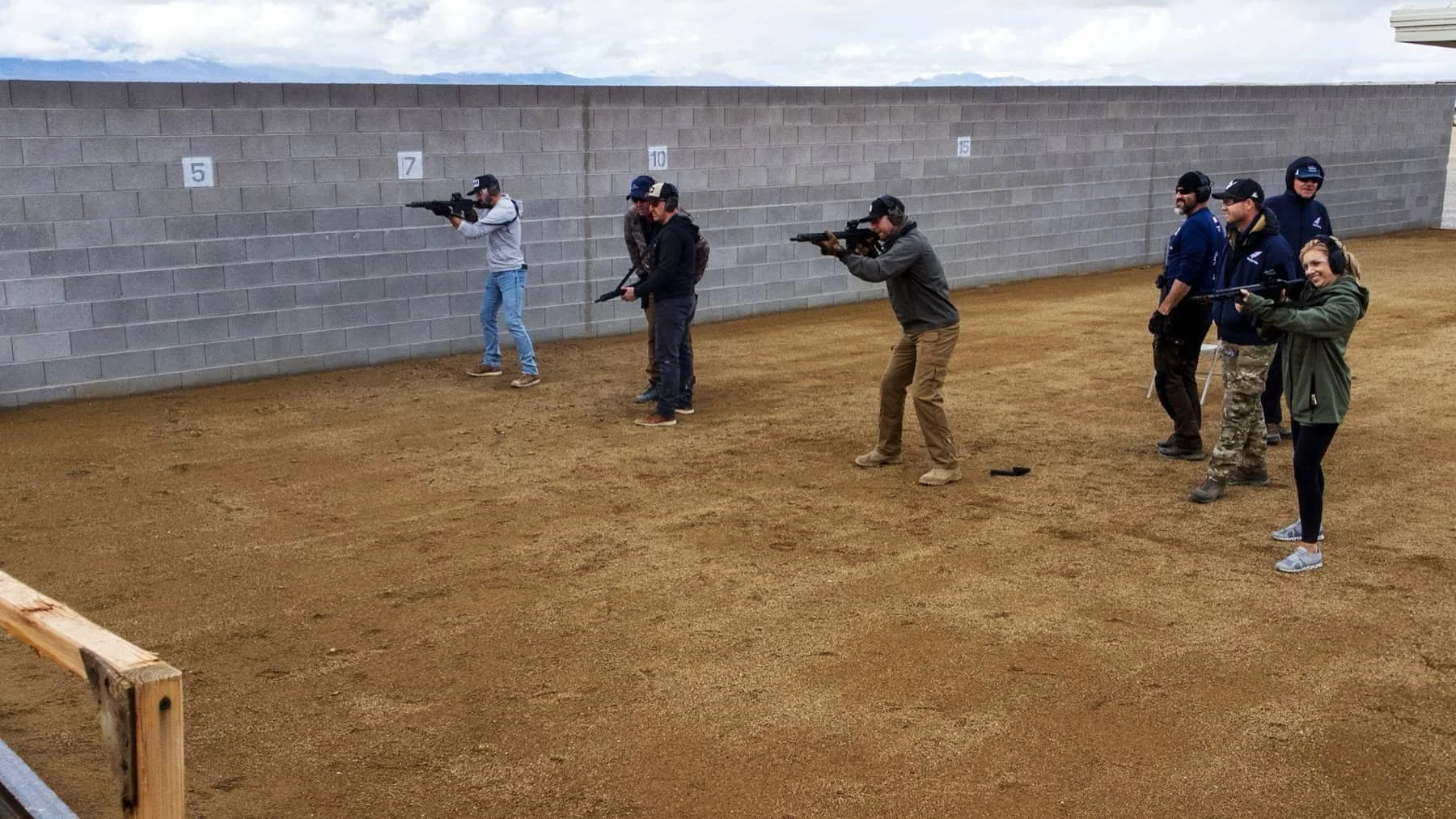 Group of people at a firing range practicing shooting with rifles, standing in front of a gray cinder block wall with numbered targets, on a dirt ground.