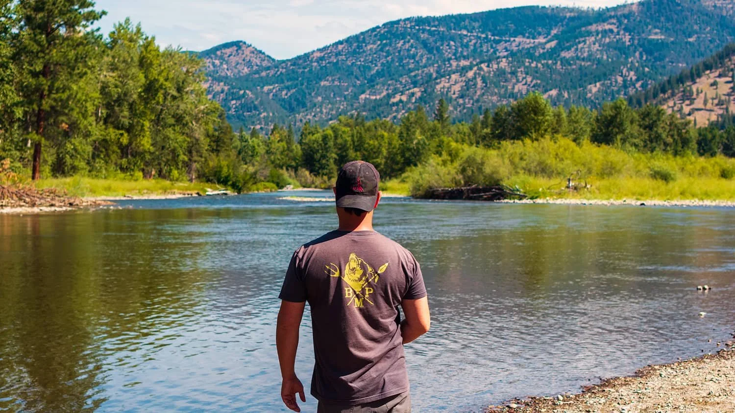 A person in a gray t-shirt and black cap standing on a rocky riverbank, looking at a river flowing through a green landscape with trees and mountains in the background.