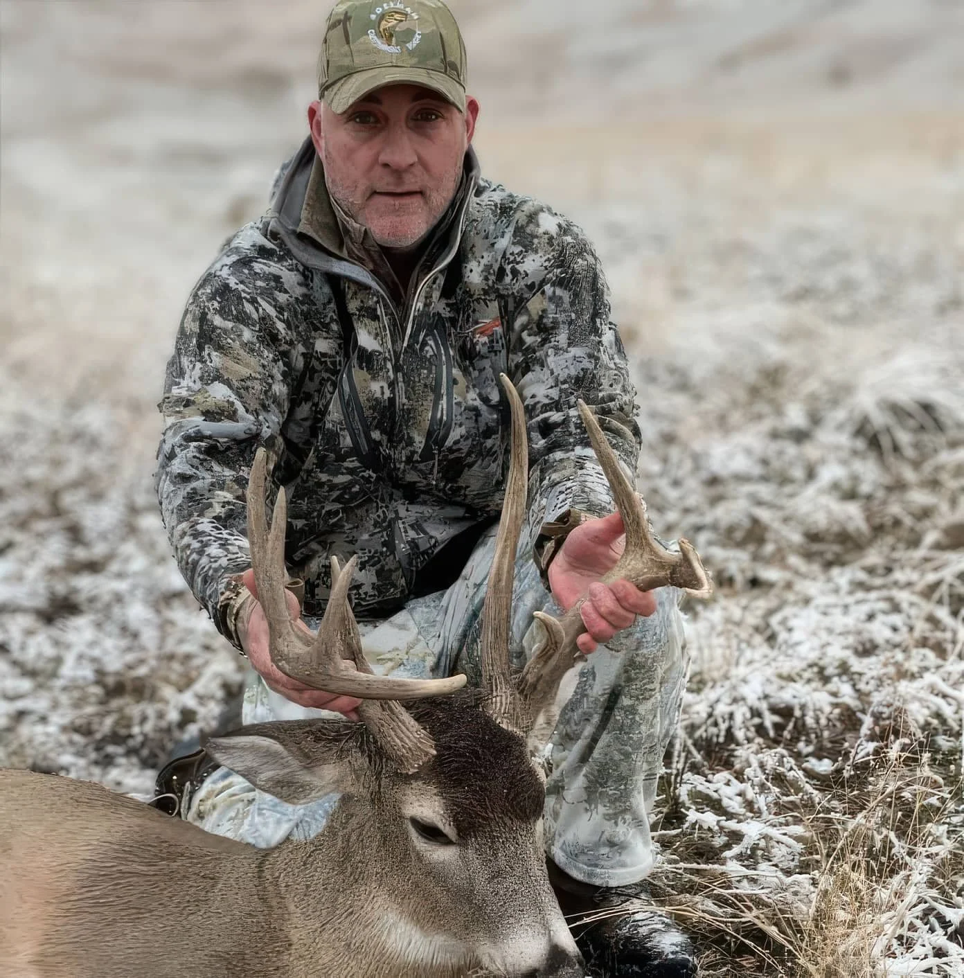 Man in camouflage holding a deer antler outdoor in a snowy landscape.