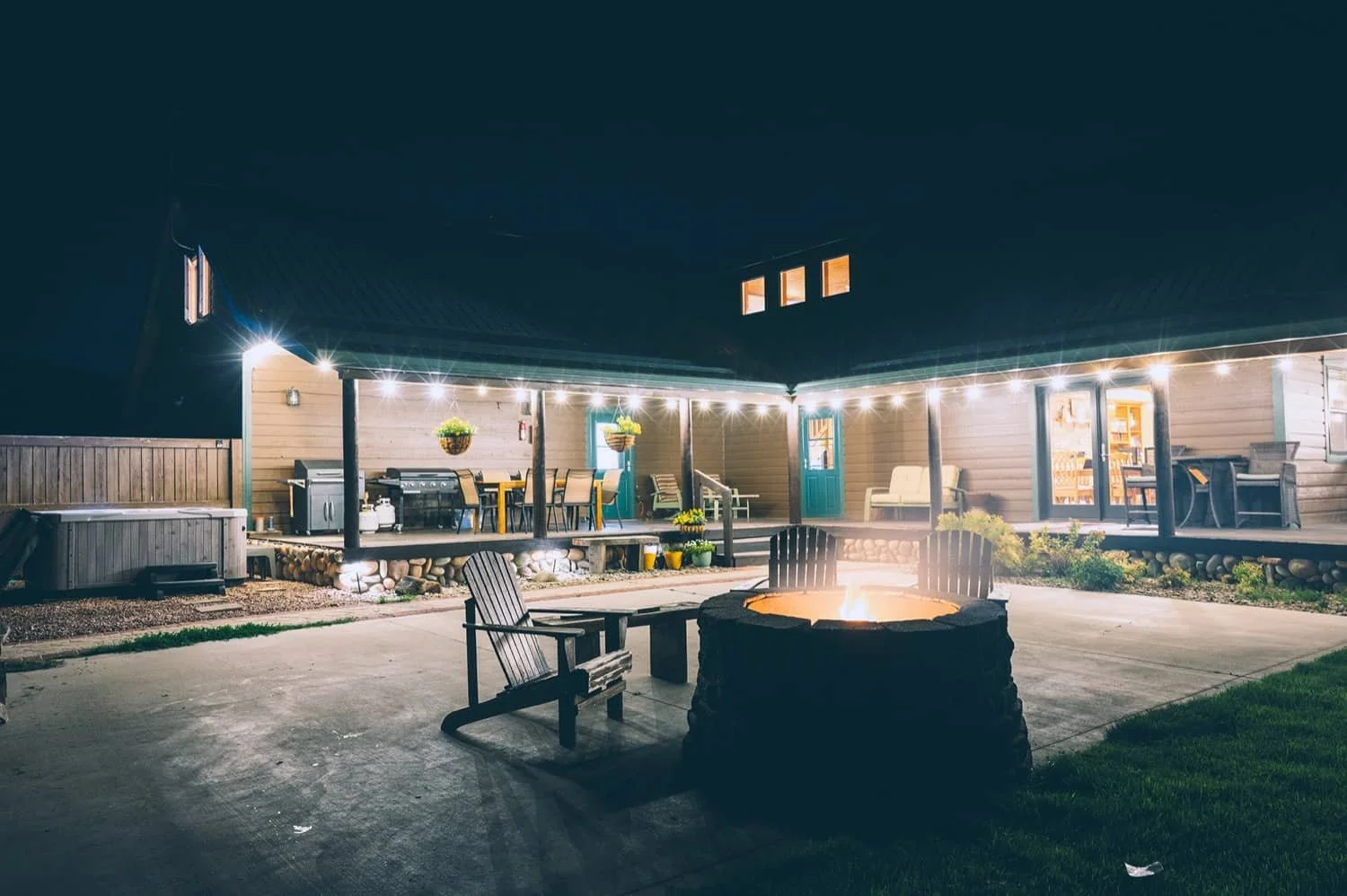 Nighttime backyard patio with string lights, fire pit, Adirondack chairs, and a covered deck with outdoor furniture, plants, and a grill.