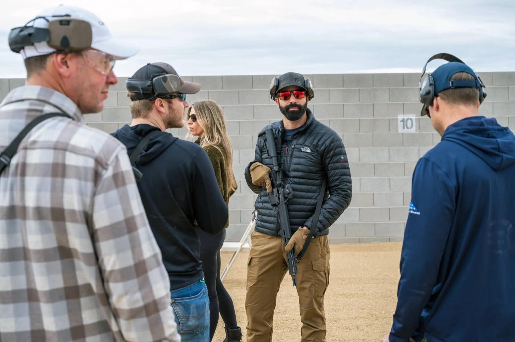 Group of people at an outdoor shooting range, all wearing hearing protection and casual clothing. One man in the center holds a rifle and wears sunglasses and gloves, with a concrete wall and sky in the background.