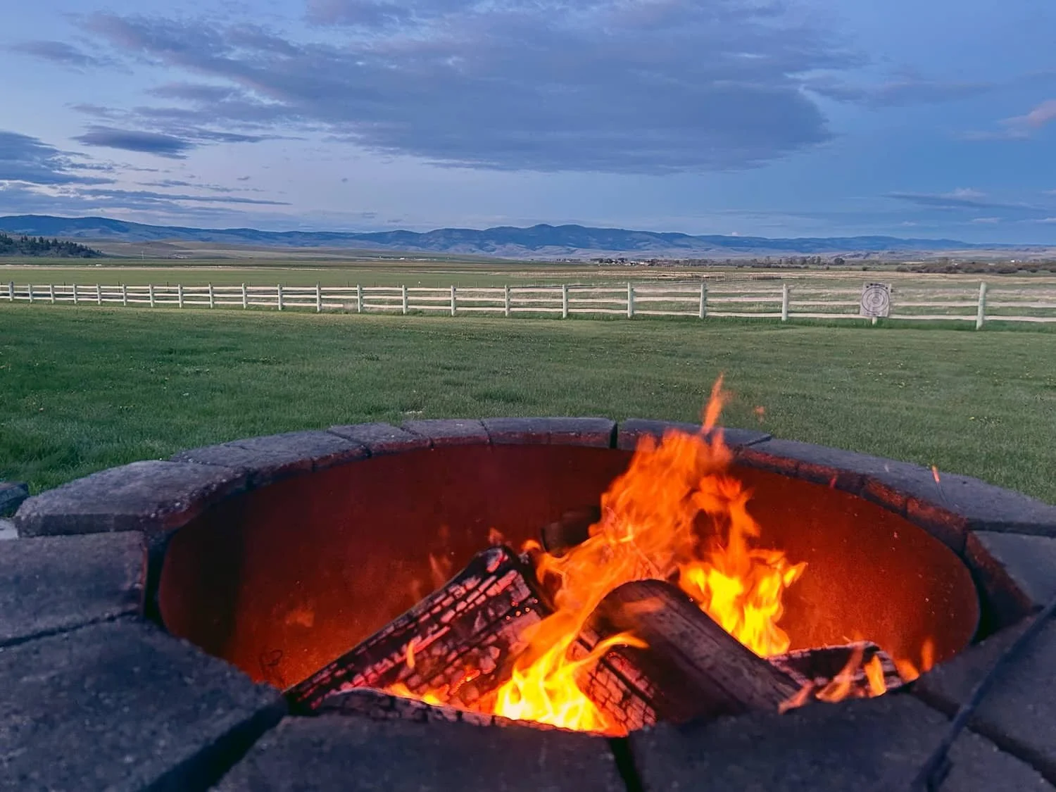 A fire pit with burning logs on a grassy lawn, with a white fence, open fields, and mountains in the distance under a sky with clouds.