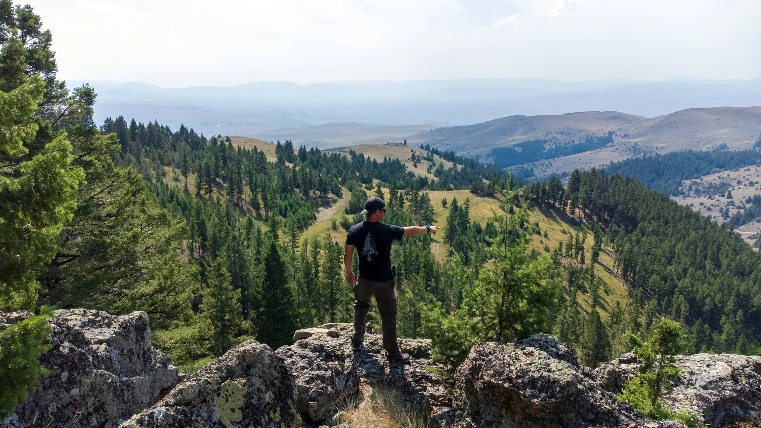 A person standing on rocks in a mountainous forest, pointing towards the distance with a backdrop of green hills and a wide valley.