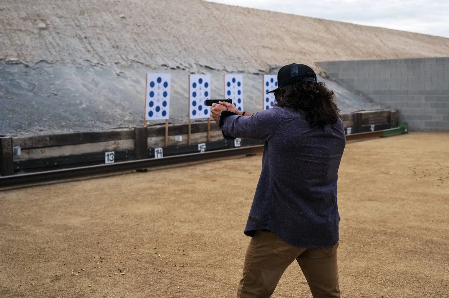 A person with long curly hair, wearing a black cap, shooting at a target range with a handgun, with silhouette targets featuring blue dots in the background.