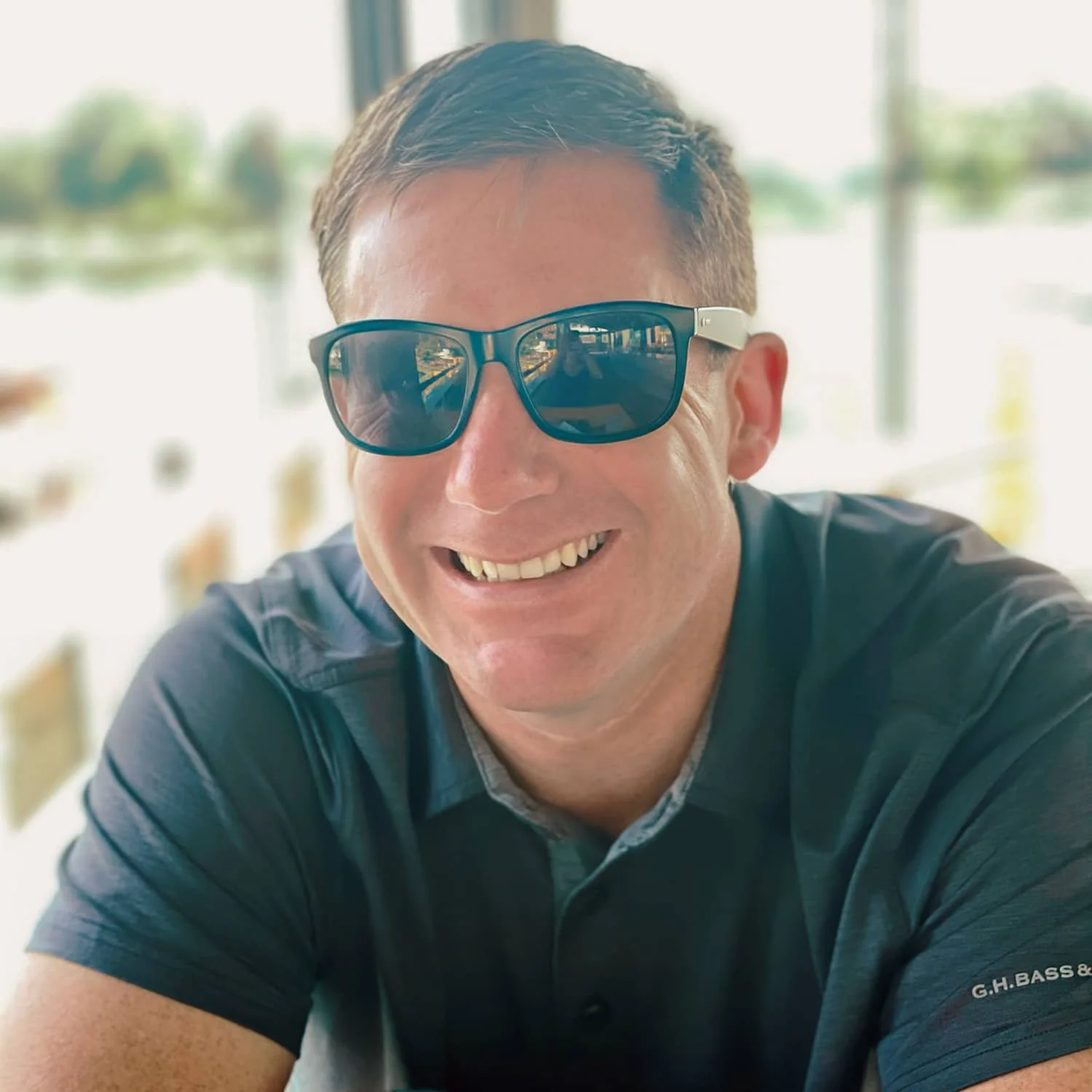 Smiling man wearing sunglasses and a dark polo shirt, sitting at a table near a window with a view of a lake or river in the background.