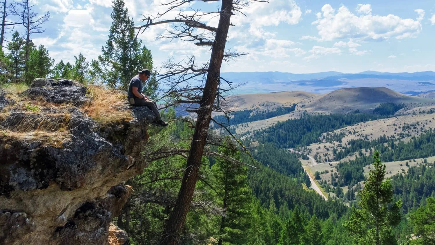 A person sitting on the edge of a rocky cliff overlooking a forested valley and rolling hills under a partly cloudy sky during daytime.