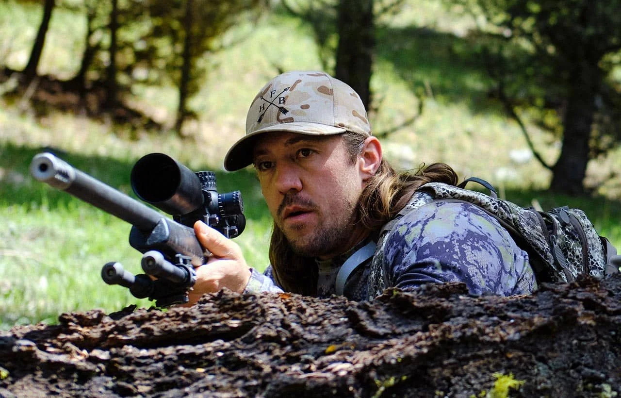 A man in camouflage military attire with a cap and backpack aiming a sniper rifle in a forest.