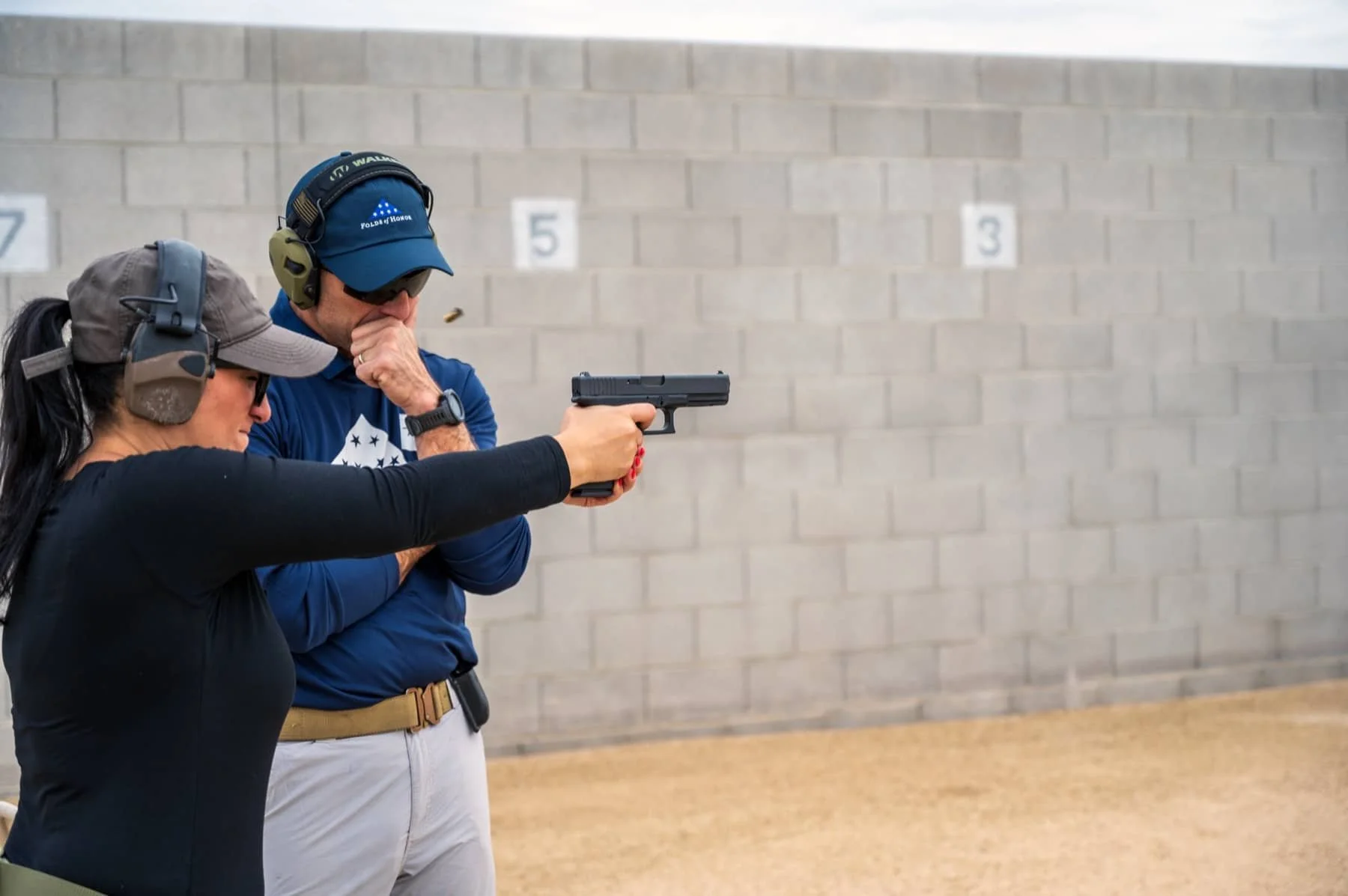 A woman practicing firearm shooting with a woman instructor at an indoor shooting range, with targets and target numbers on the wall behind them.
