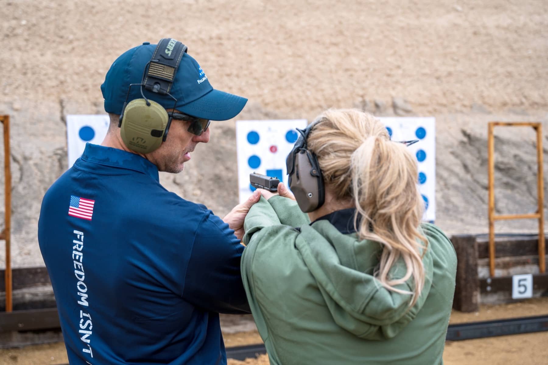 Two people, a man and a woman, at a shooting range, with the man helping the woman aim a handgun. Both are wearing ear protection and the man is wearing a blue jacket with the American flag and the words 'U.S. FREEDOM' on the back. The background features shooting targets with blue and red dots and a beige wall.