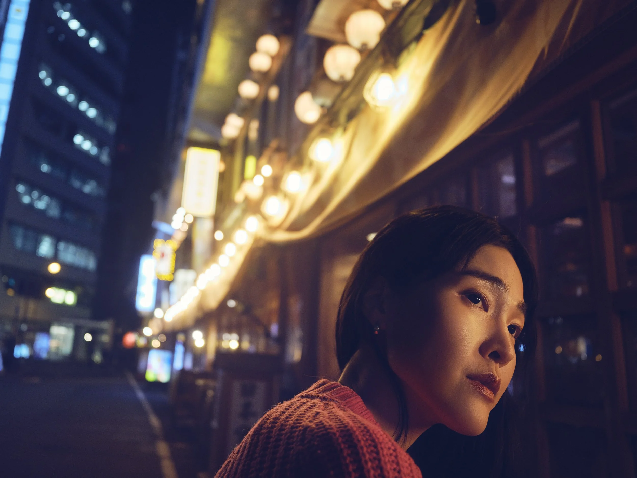 A woman with dark hair and earrings at night in an urban setting illuminated by streetlights and neon signs. Japan, Tokyo