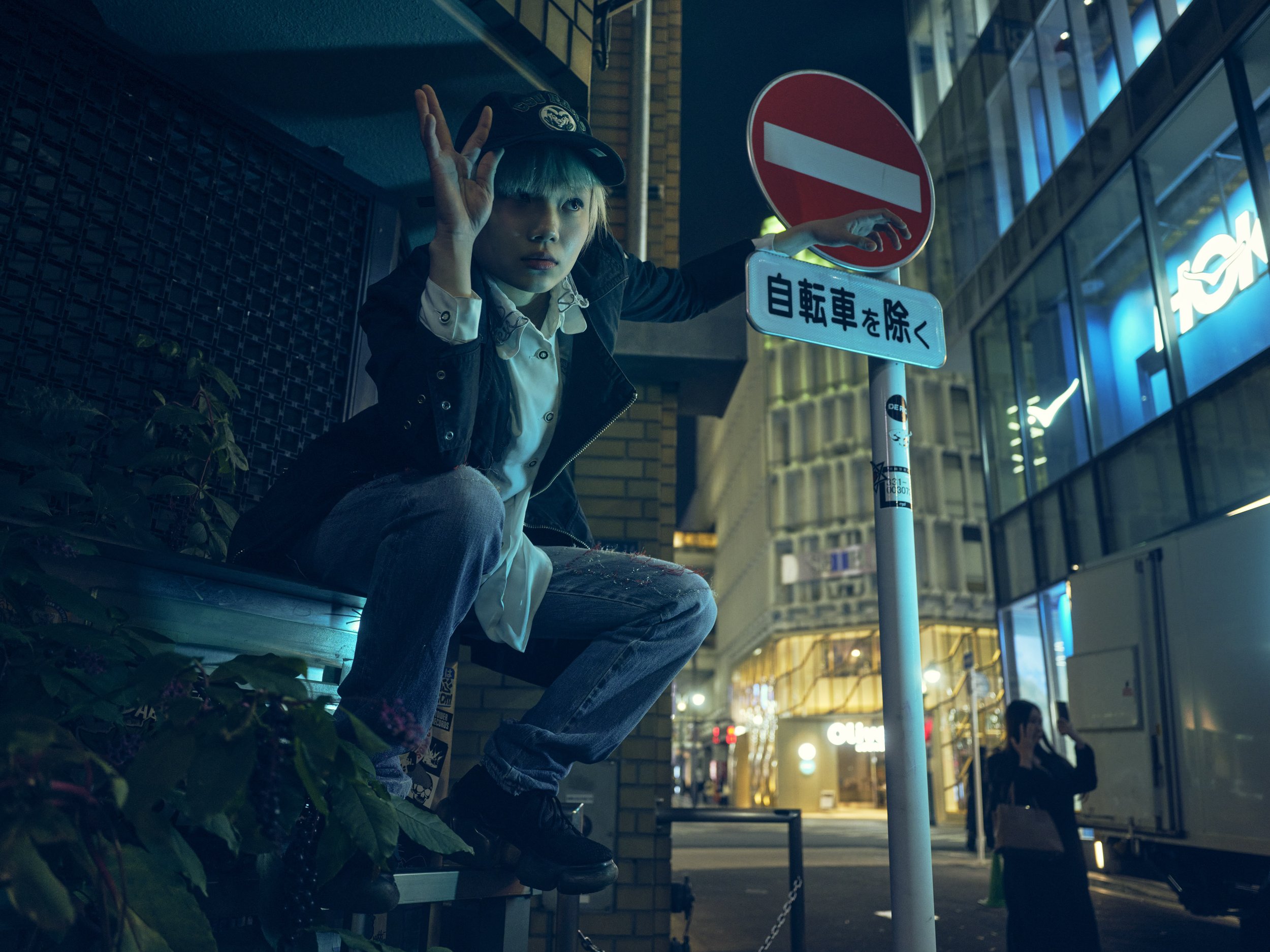 A young person with a cap crouches on a ledge at night, holding a 'no entry' sign, in an urban area with illuminated shops and a person talking on a phone in the background.