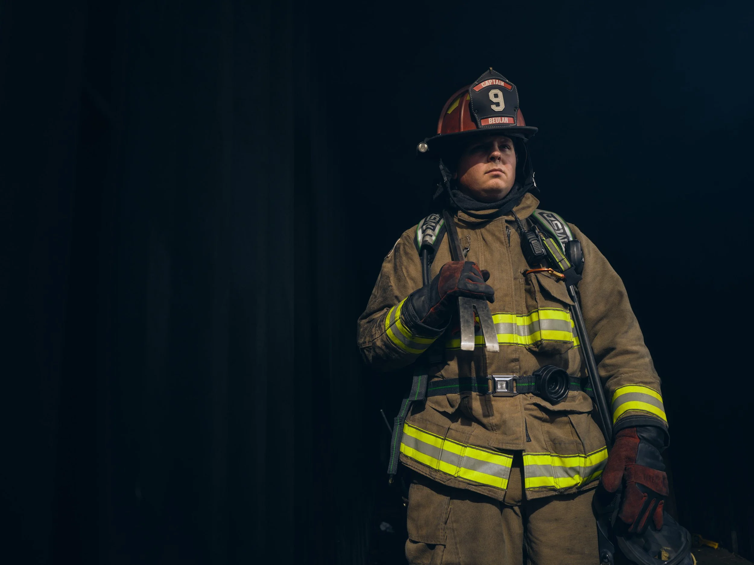 Firefighter in uniform holding a helmet, standing in a dark environment, looking serious.