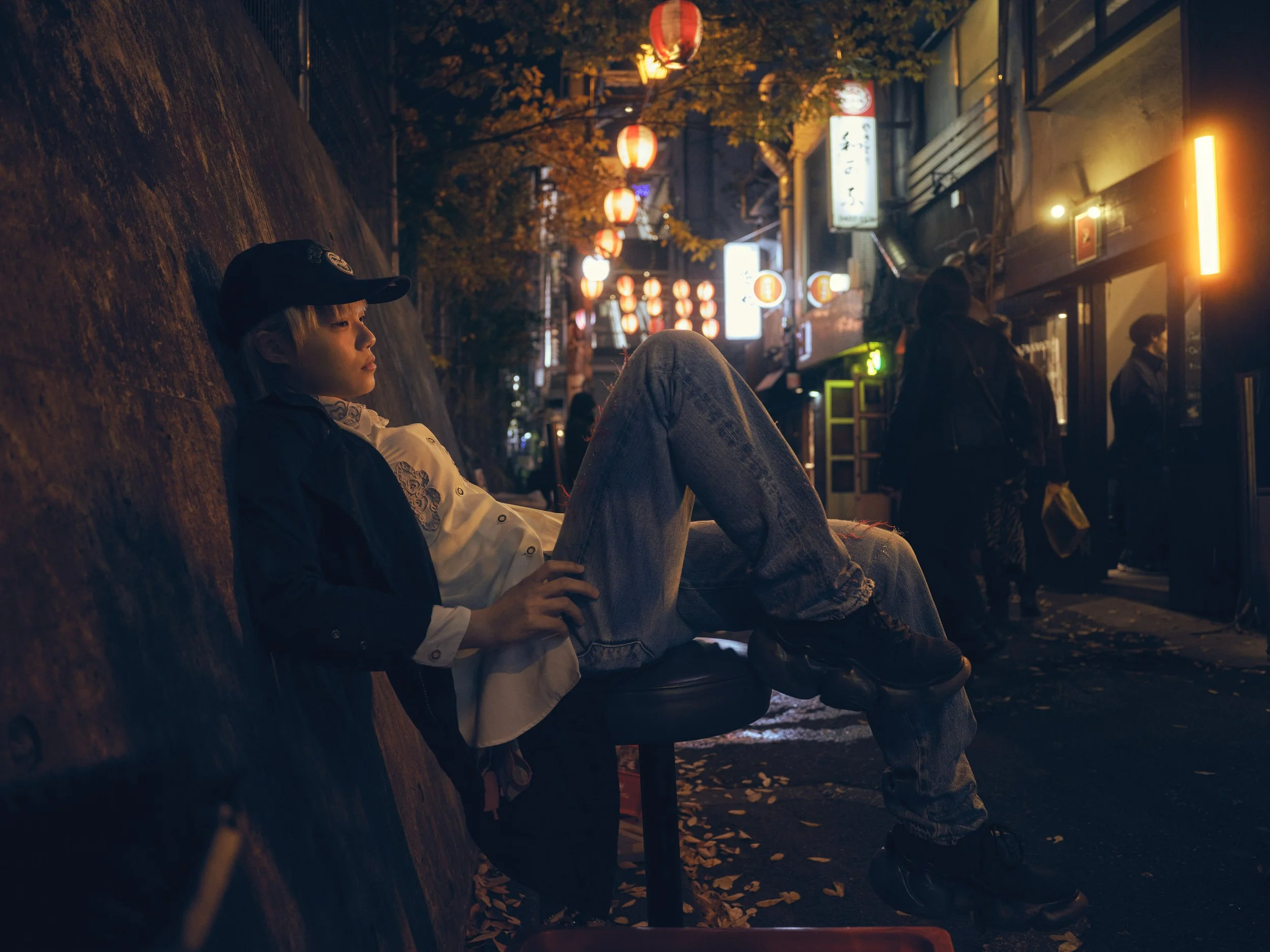 A young person wearing a black cap, white shirt, and black jacket, sitting sideways on a stool against a sloped wall at night in an urban setting. The street is decorated with hanging red lanterns, and there are passersby and illuminated signs in the