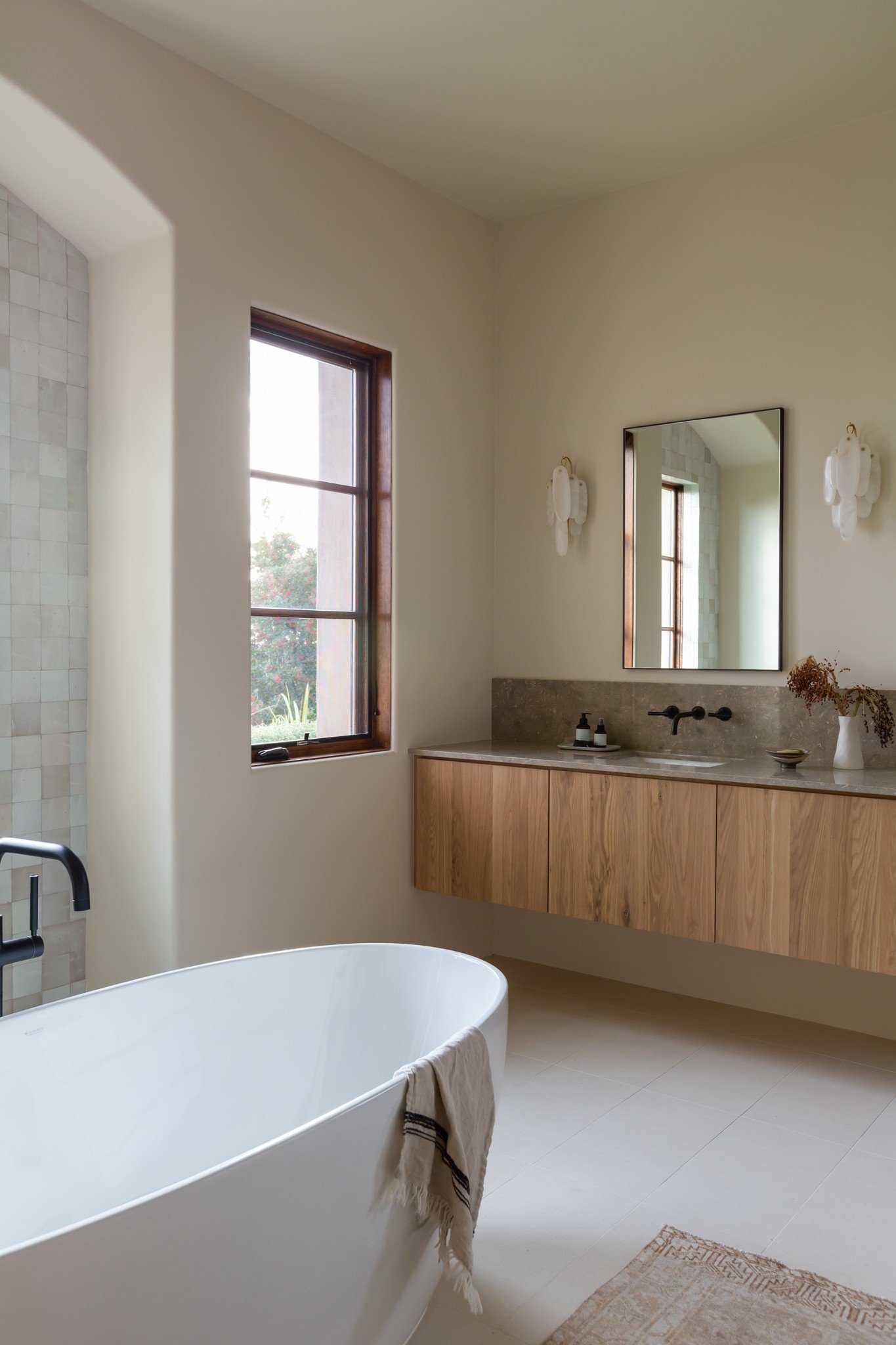 Modern luxury bathroom with white soaking tub, black accent windows, wooden floating cabinets and sink.