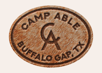 Wooden oval badge with 'Camp Able' and 'Buffalo Gap, TX' engraved around the edge, and a central logo with the letters C and A intertwined.