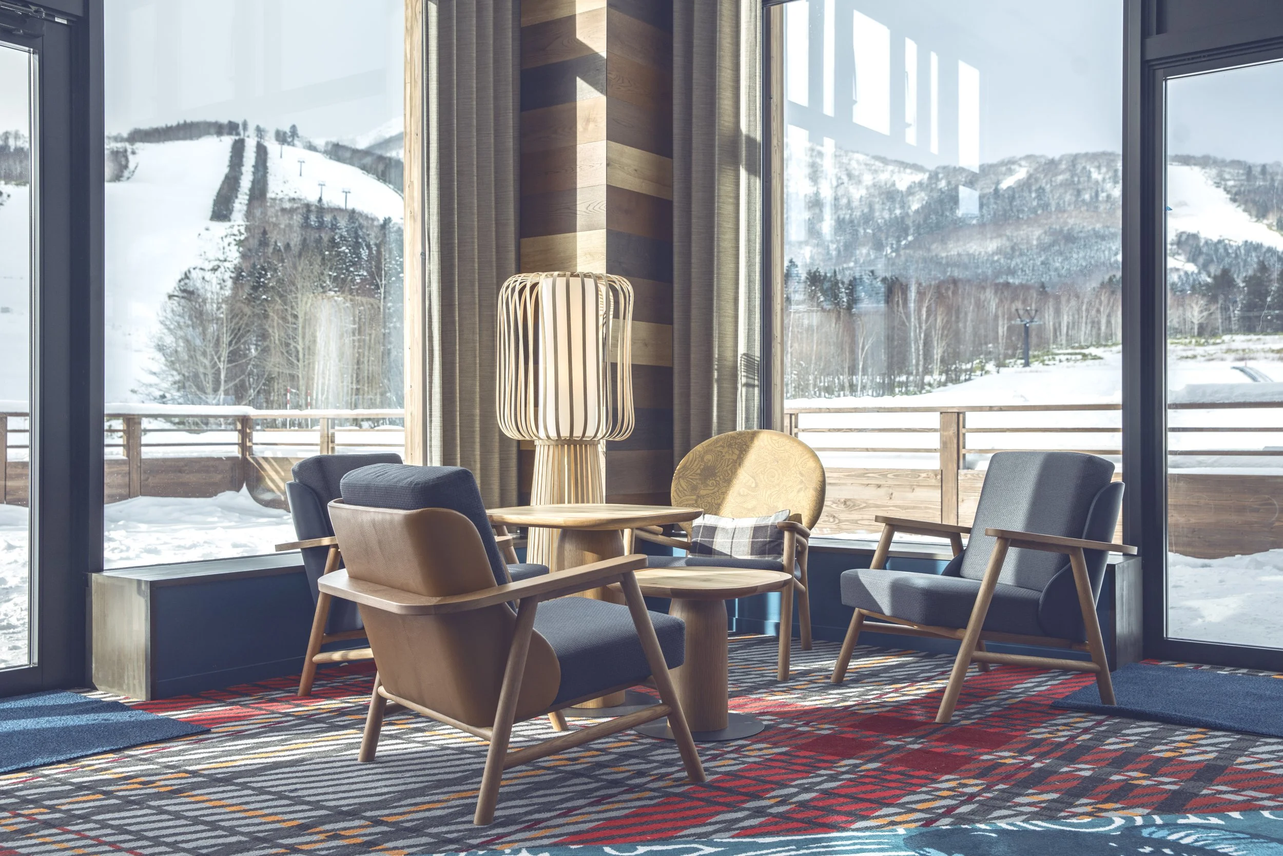 Interior of a cozy lounge area with five chairs around a small wooden table, large windows showing snowy mountains and trees outside, and a striped rug on the floor.