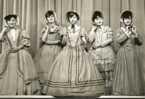 Five children in old-fashioned dresses and bonnets pose in front of a curtain, smiling and holding their hats. Black and white photo. Part of a drama production in the 1960s