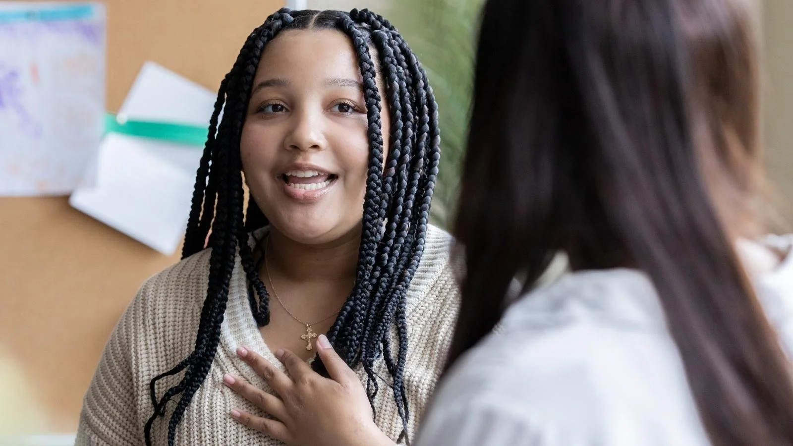 A smiling student with braided hair speaks animatedly to another person in a school setting, hand on chest, capturing the warmth of personal storytelling and authentic voice.