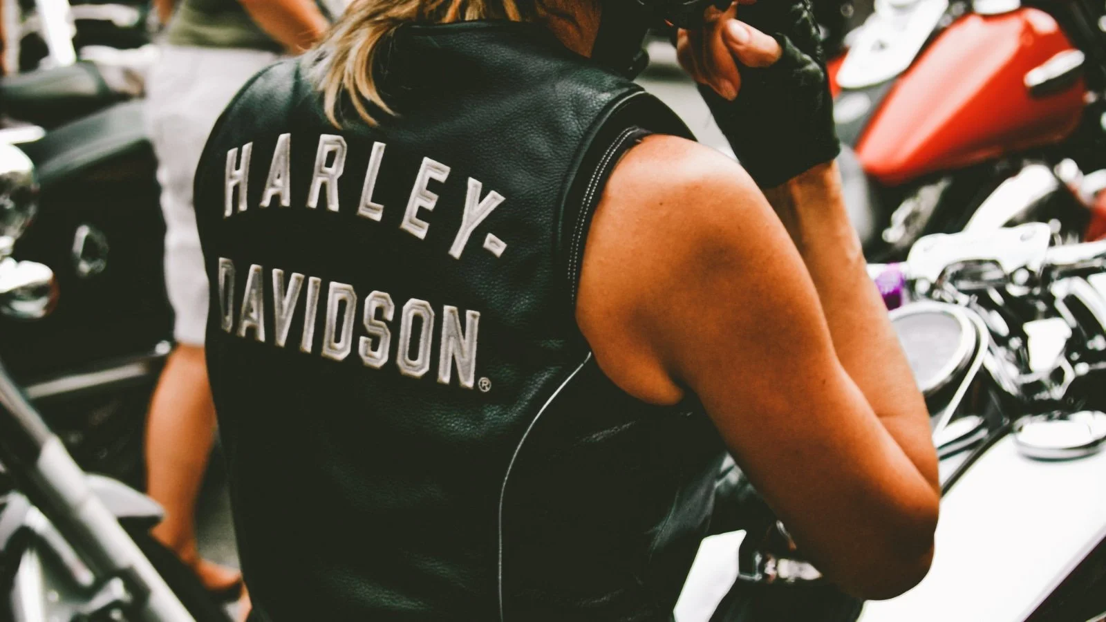 A person wearing a sleeveless Harley-Davidson leather vest sits on a motorcycle among other bikes.