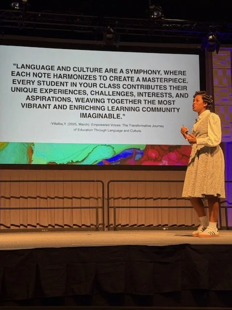 Young woman in a beige dress speaks on stage beside a large screen displaying a quote about language and culture.