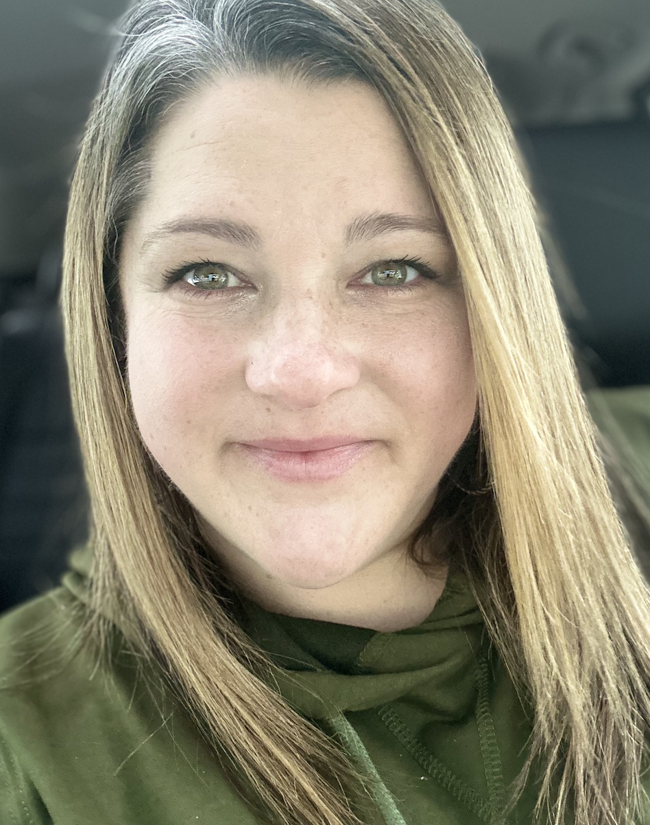Close-up portrait of a woman with shoulder-length light brown hair, green eyes, and light skin, smiling softly, wearing a green jacket.