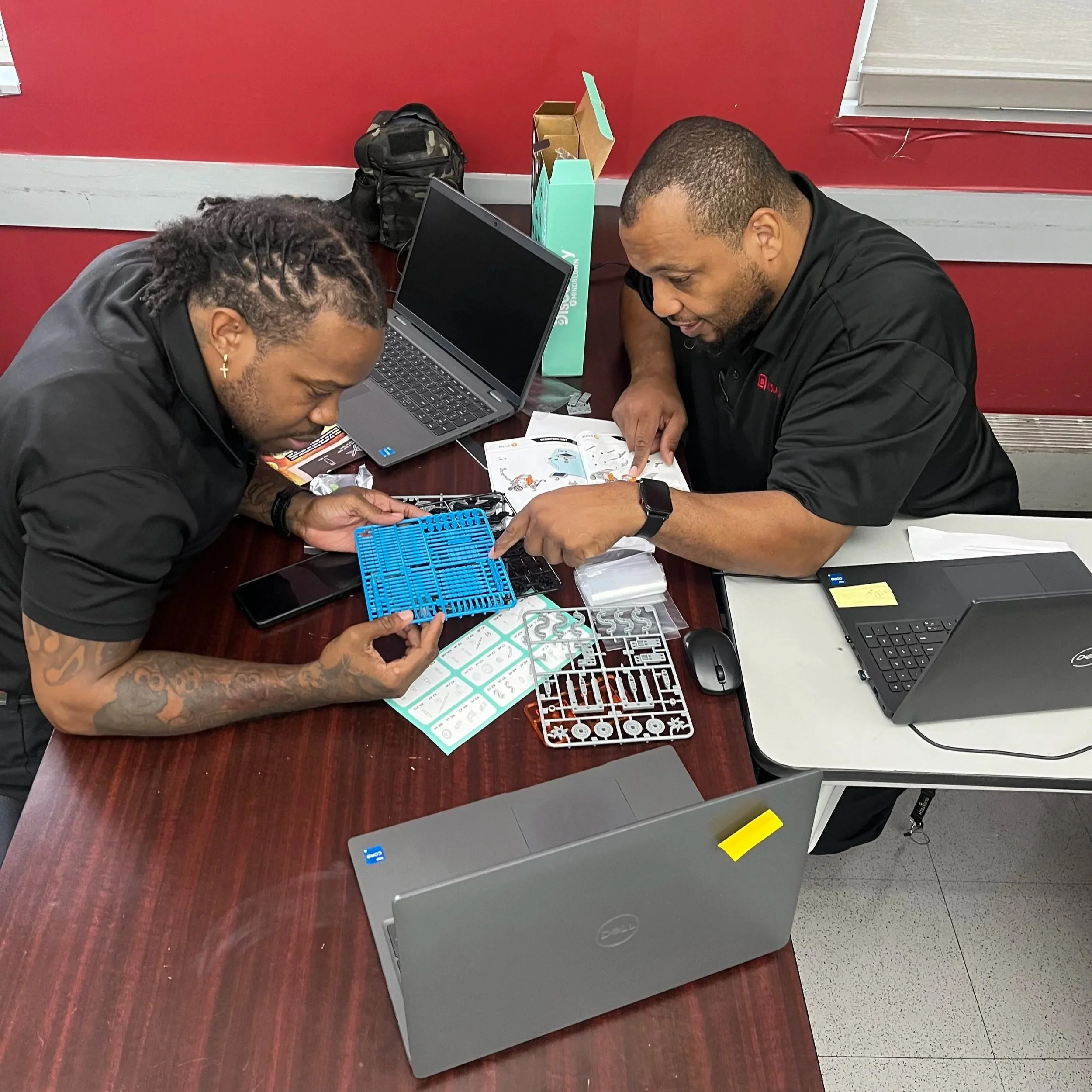 Two men are sitting at a wooden table, working on a model kit with plastic parts and instruction sheets. One man has tattoos on his arms and is holding a blue tool, while the other is pointing at the instruction sheet. There are two laptops, a backpack, a green box, and a small black box on the table in a room with a red wall and a window.