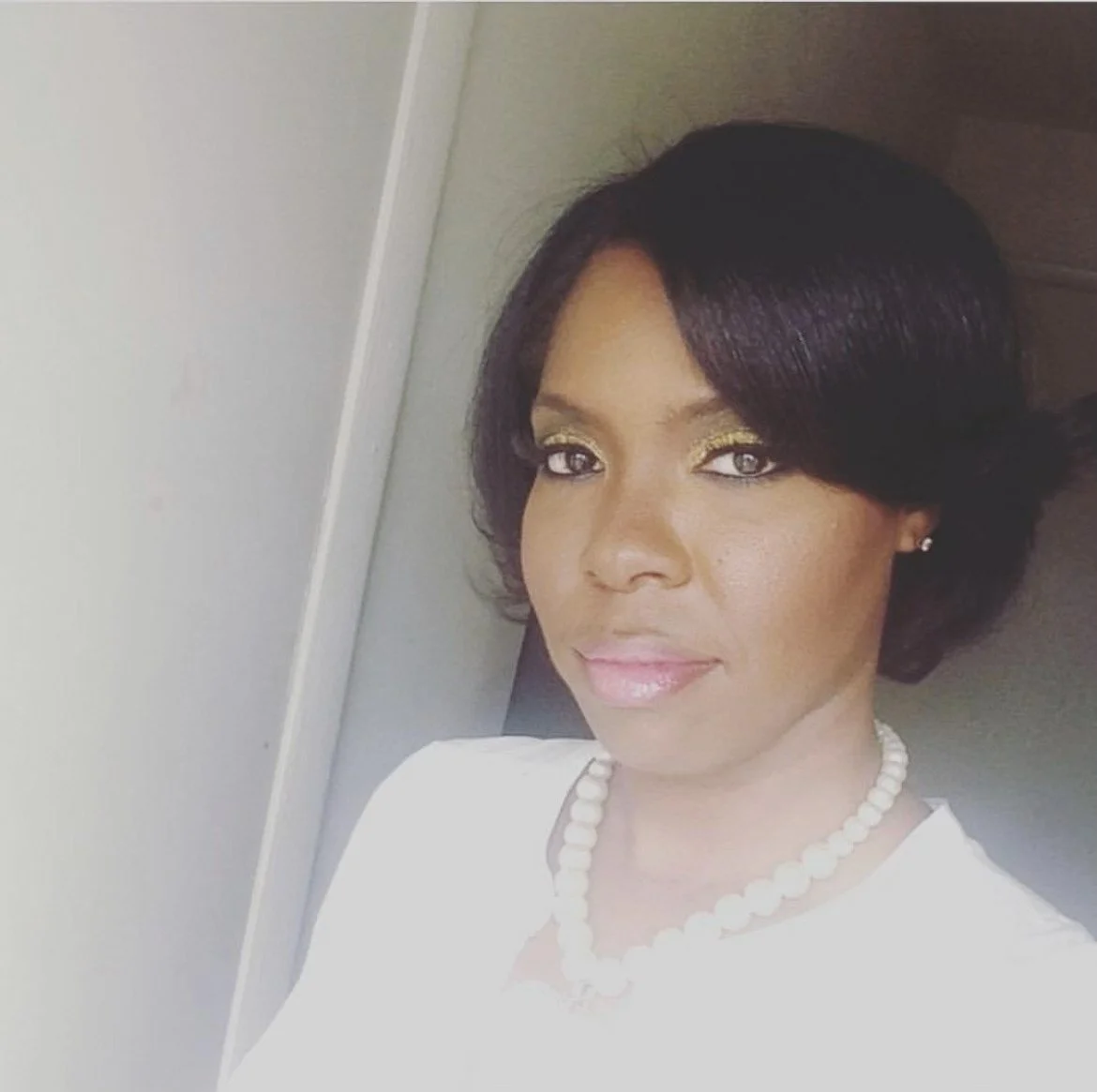 A woman with short black hair, wearing gold eyeshadow, pink lipstick, pearl necklace, and earrings, looking at the camera.