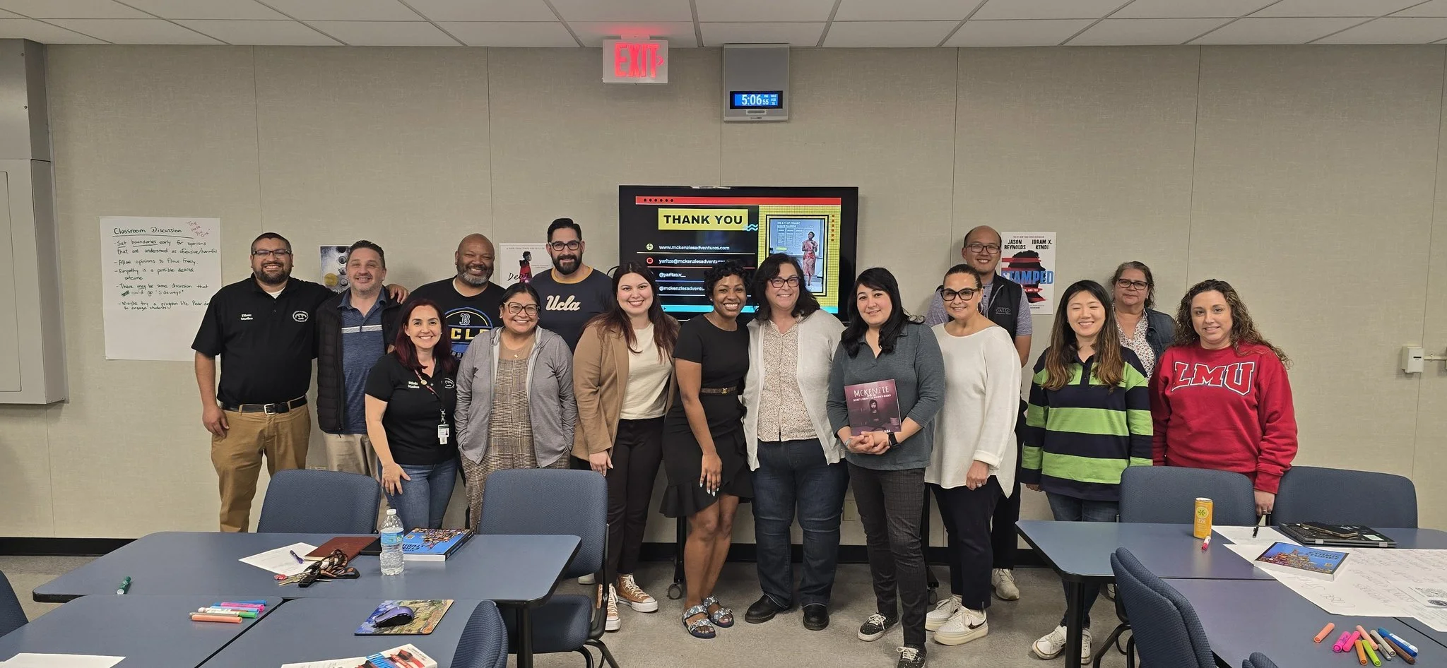 Group of fifteen diverse people standing together in a classroom or conference room, smiling for a photo. The room has a digital clock showing 5:06, a large screen displaying a thank you message and social media information, and whiteboards on the walls.