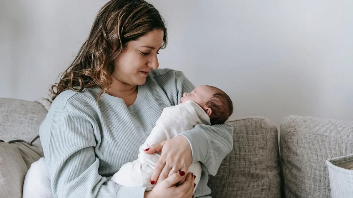 A mother smiling gently while holding her sleeping newborn baby swaddled in a white blanket on a living room couch.