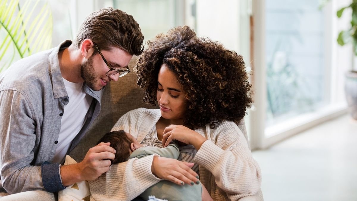 A mother with curly hair and a father wearing glasses sitting together, smiling down lovingly at their newborn baby