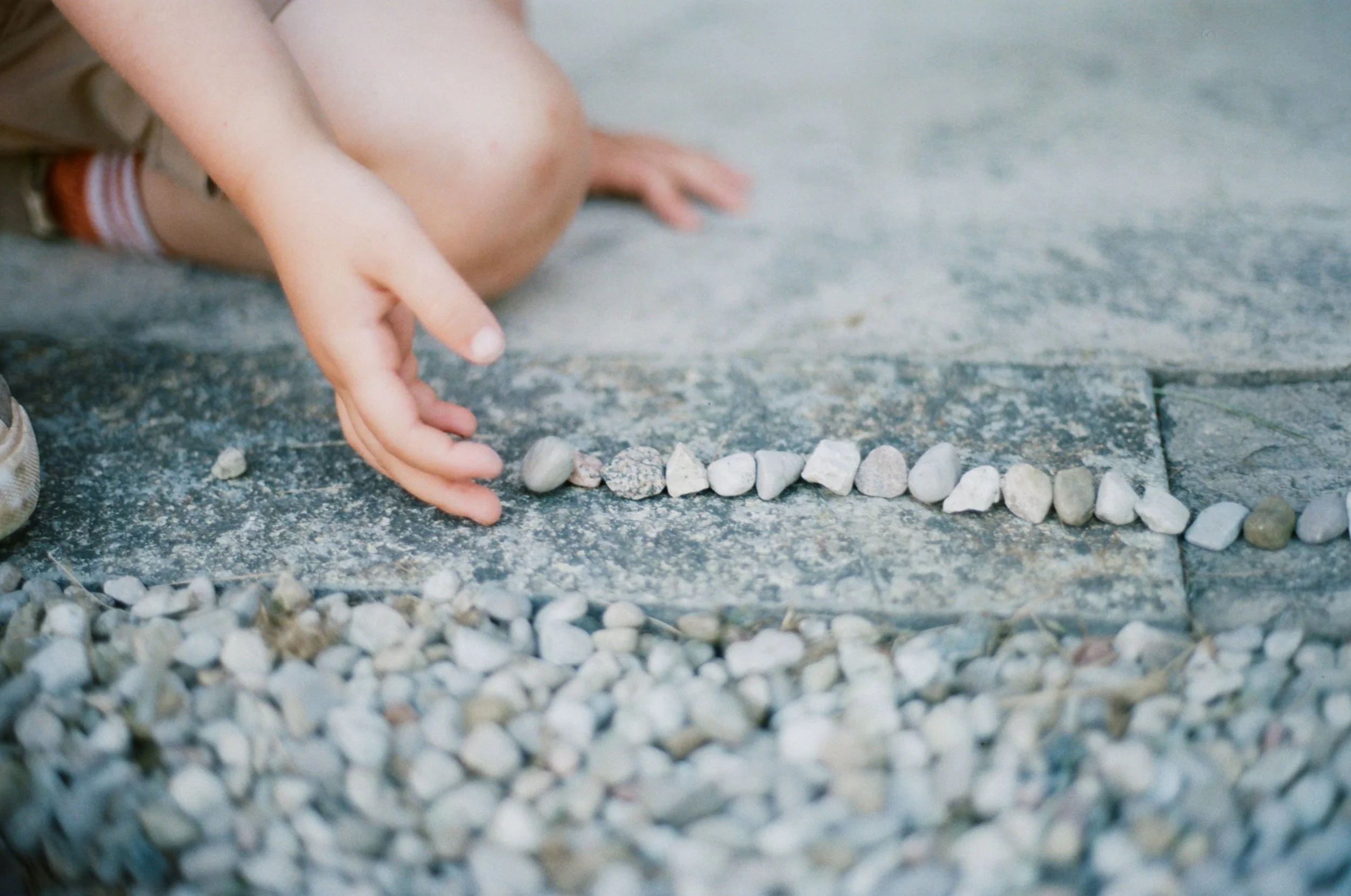 A child wearing shorts and sneakers is sitting on the ground, arranging small rocks in a line on concrete pavement.